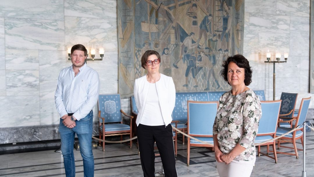 Tre personer står innendørs foran et stort billedvev, med stoler og stearinlys i bakgrunnen.