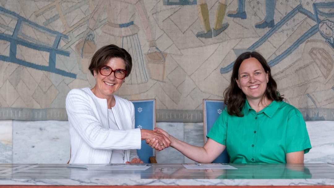 To kvinner sitter ved et bord, smiler og håndhilser, foran et billedvev. Man bruker briller og hvit jakke; den andre har på seg en grønn skjorte.