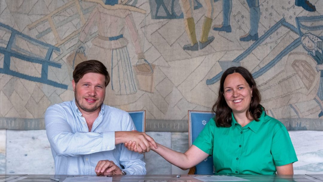 To personer sitter ved et bord, håndhilser og smiler. I bakgrunnen er det et dekorativt veggteppe. Personen til venstre har på seg en lyseblå skjorte; personen til høyre har på seg en grønn skjorte.