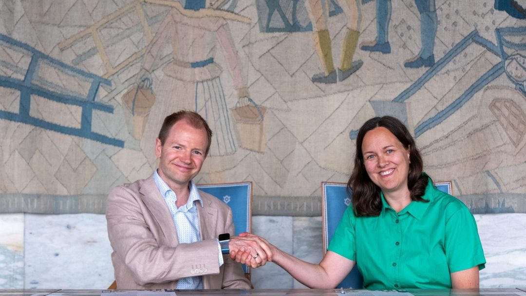 En mann i beige dress og en kvinne i en grønn skjorte håndhilser mens de sitter ved et bord med et fargerikt veggmaleri i bakgrunnen.