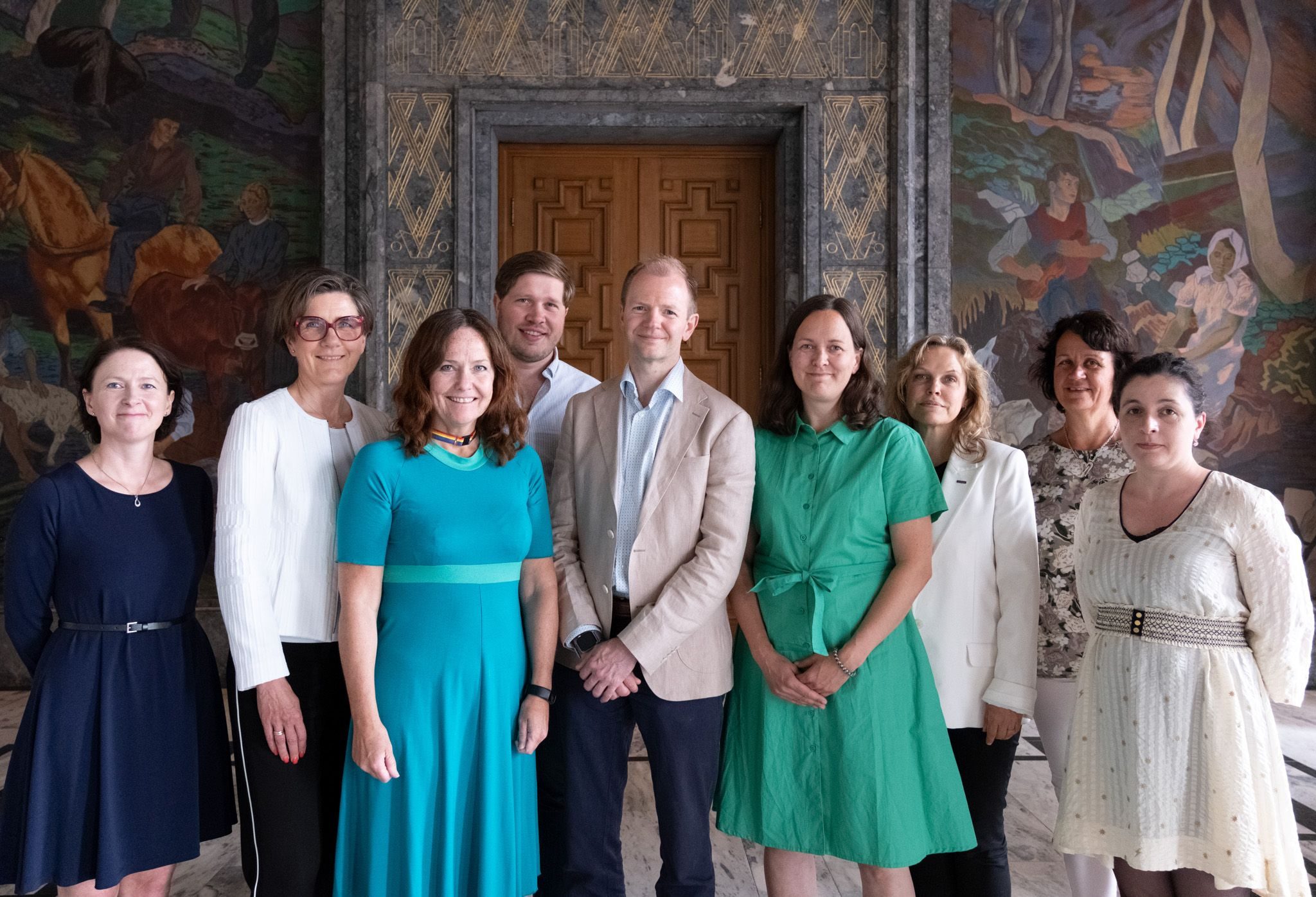 En gruppe på ni personer som står innendørs foran en dekorativ vegg og smiler til kameraet.