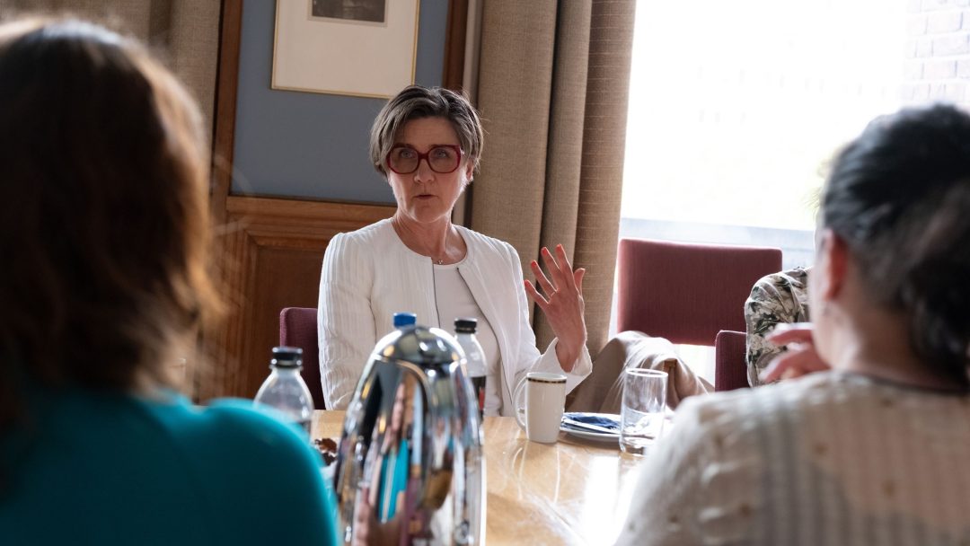 En person i hvitt snakker ved et bord med andre, omgitt av vannflasker og en kaffekanne.