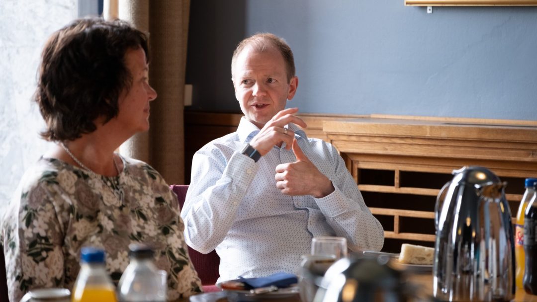 To personer sitter ved et bord med drinker, engasjert i en samtale. Bordet har flasker, termos og glass.