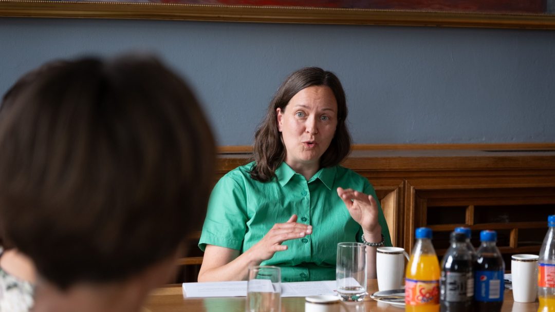 En kvinne i en grønn skjorte snakker animert på et møte, sittende ved et bord med drinker og papirer.