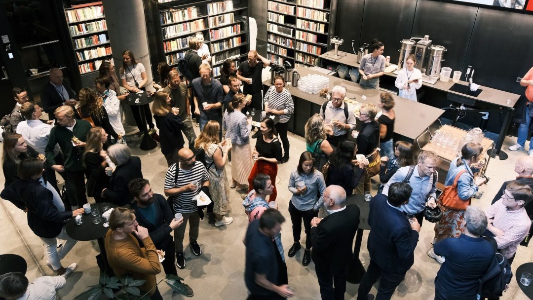 En stor gruppe mennesker som sosialiserer seg på et bibliotekarrangement, omgitt av bokhyller og forfriskningsbord, hvor noen sitter og andre står.