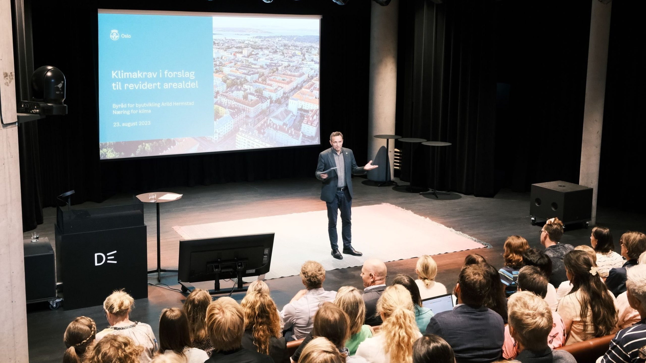 En mann i dress holder en presentasjon for et sittende publikum i en moderne, svakt opplyst sal. En stor skjerm viser et lysbilde på et fremmedspråk om klimainitiativer.