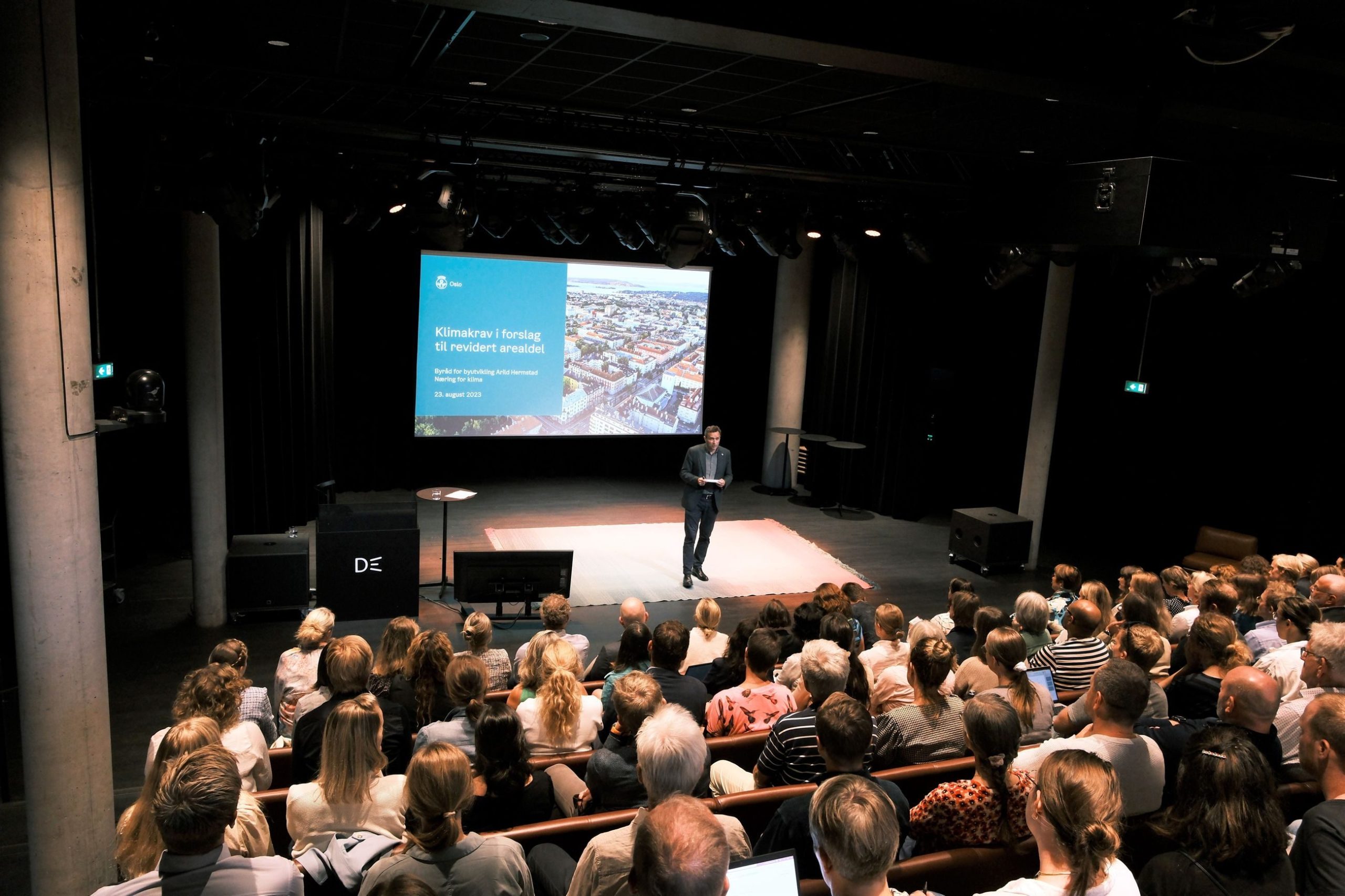 En mann står på scenen og presenterer for et publikum i et teater. En skjerm viser tekst og et bybilde fra luften.