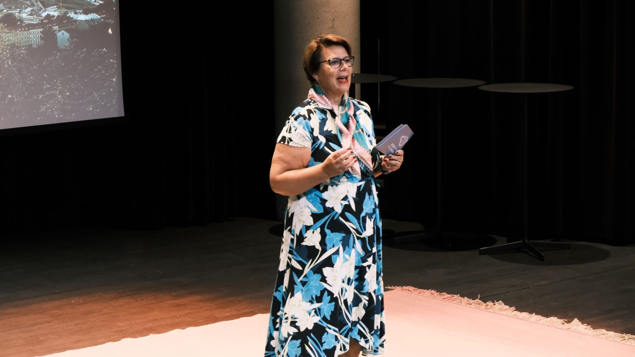 En kvinne i en blomsterkjole snakker på scenen og holder et hefte. En presentasjonsskjerm er synlig i bakgrunnen.
