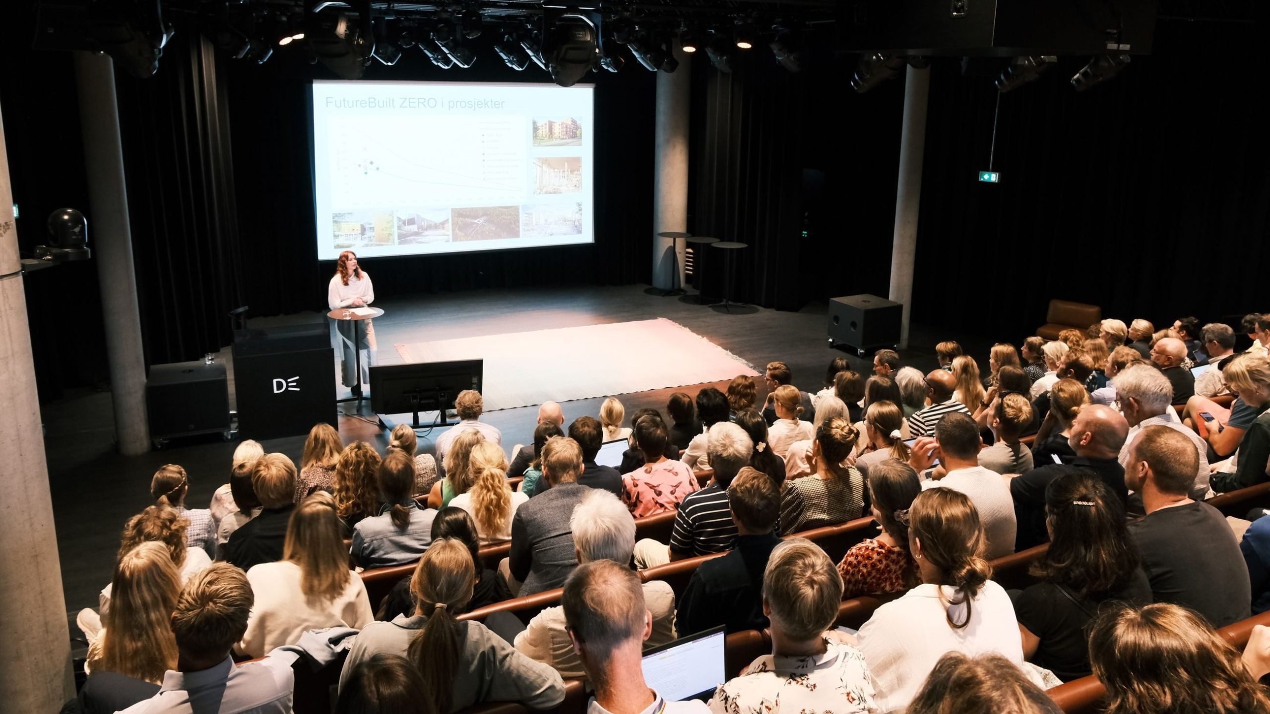 En foredragsholder står på et podium foran et publikum i et mørkt auditorium, og presenterer lysbilder på en skjerm med tittelen FutureBuilt ZERO.