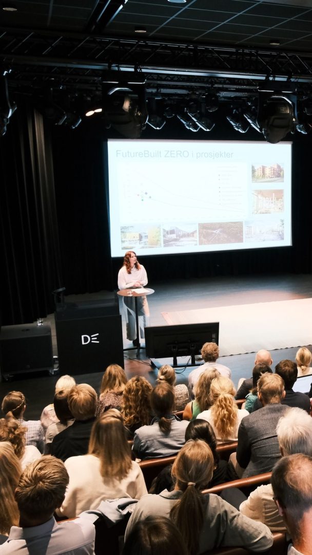 En foredragsholder står ved et podium på en scene og henvender seg til et publikum. Et presentasjonslysbilde med tittelen FutureBuilt ZERO i prosjekter vises på en skjerm bak dem.