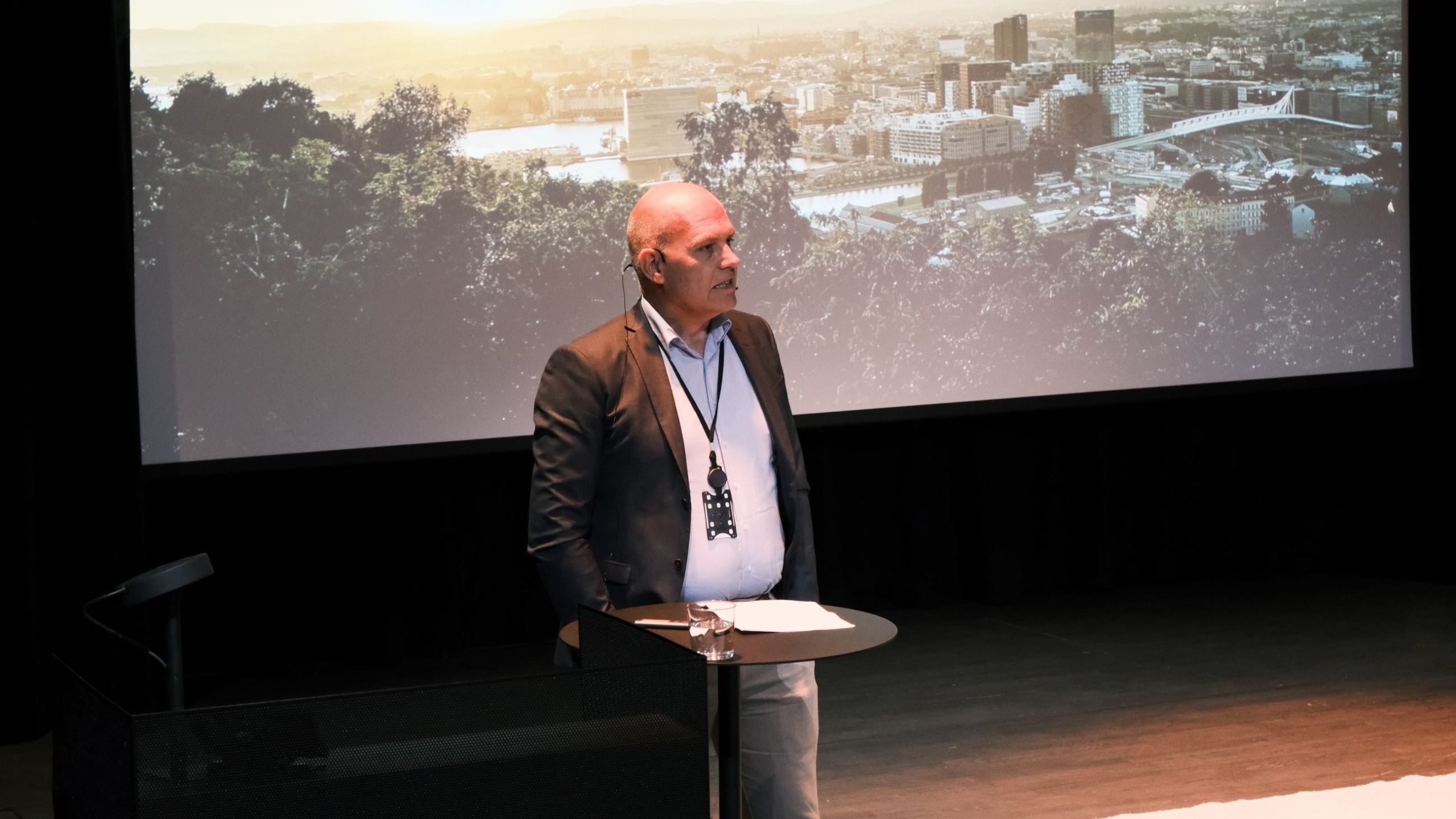 En mann står på et podium med notater, iført et headset. Bak ham er et bybilde projisert på en lerret.