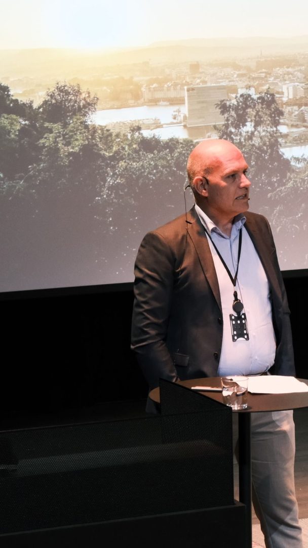 Mann som står ved en talerstol og snakker foran et publikum. Bak ham er en stor skjerm som viser et bybilde og grøntområder.