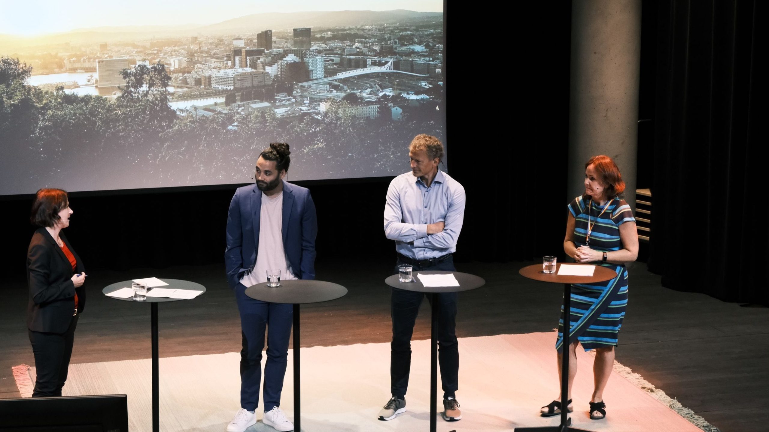Fire personer står på scenen ved høye bord, engasjert i diskusjon, med et bybilde projisert på en skjerm bak seg.