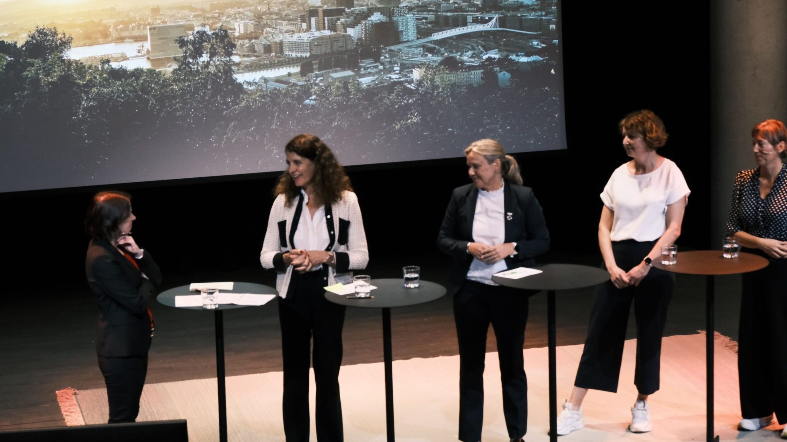 Fem personer står på en scene med høye bord og deltar i en paneldiskusjon. Et bybilde er projisert bak dem.