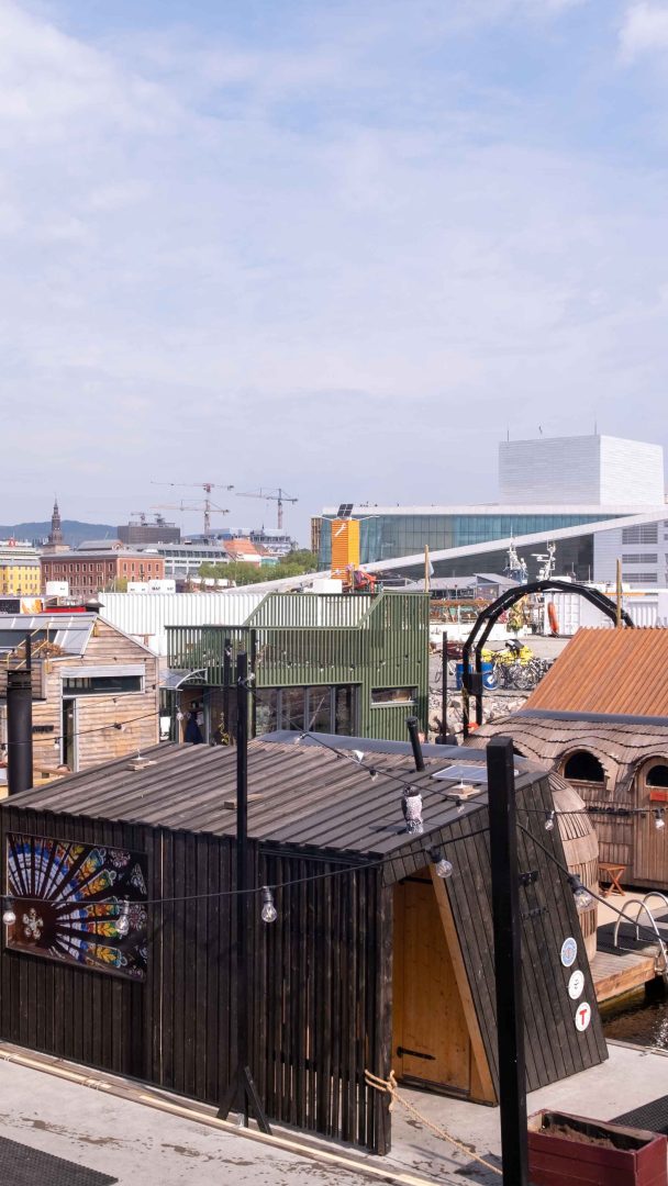 Trekonstruksjoner og hytter er i forgrunnen, og viser en rustikk sjarm, mens urbane bygninger og byggekraner reiser seg bak dem under en overskyet himmel, og tilbyr autentiske Oslotips for de som utforsker kontrasten mellom gammelt og nytt.