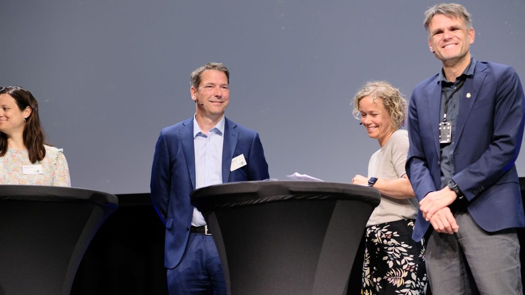 Fire personer står ved runde podier på en scene under et arrangement. To menn har på seg dress, og to kvinner er kledd i uformelle klær. En grå bakgrunn er synlig.