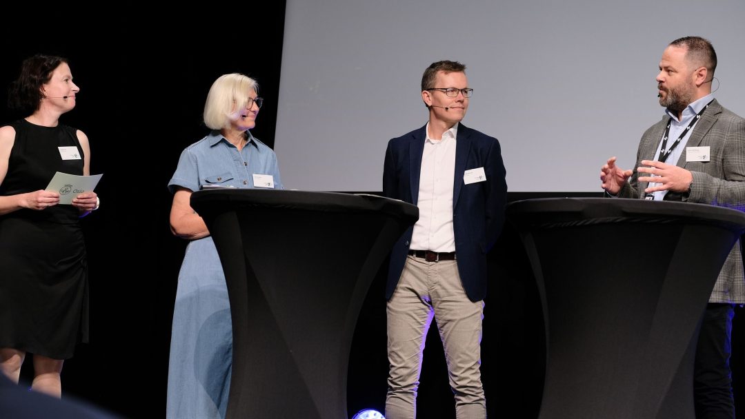 Fire personer i forretningsantrekk står og snakker rundt to høye bord under en konferanse eller panelsamtale.