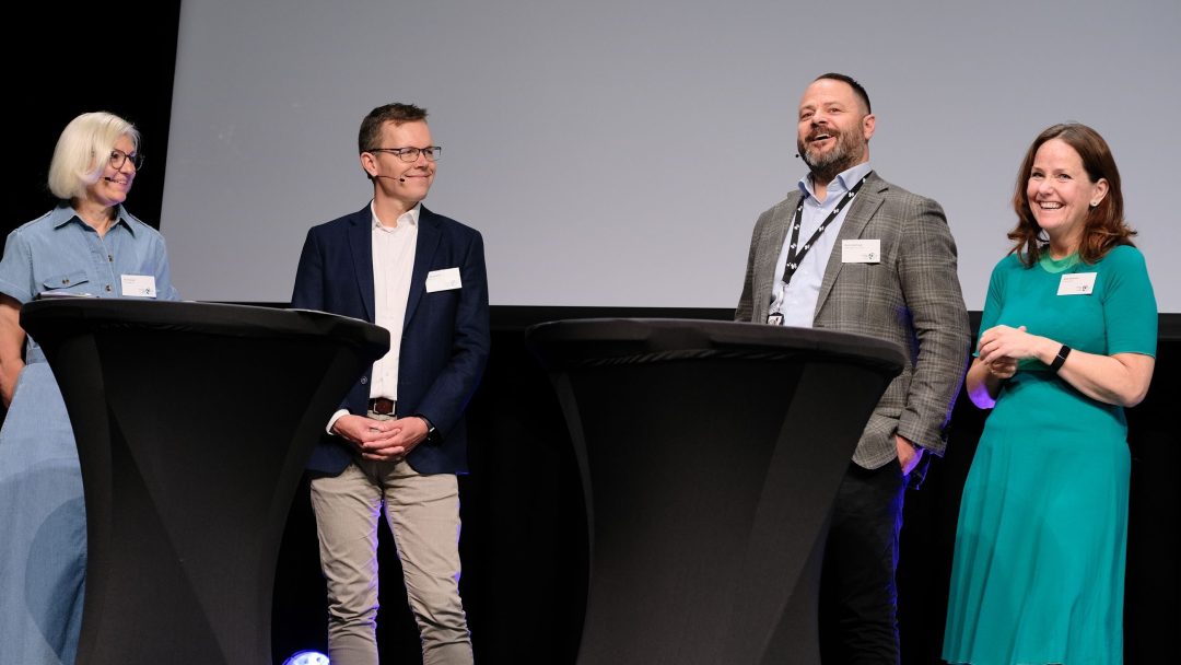 Fire personer står og snakker ved to runde bord på en scene, hver med navnelapper.