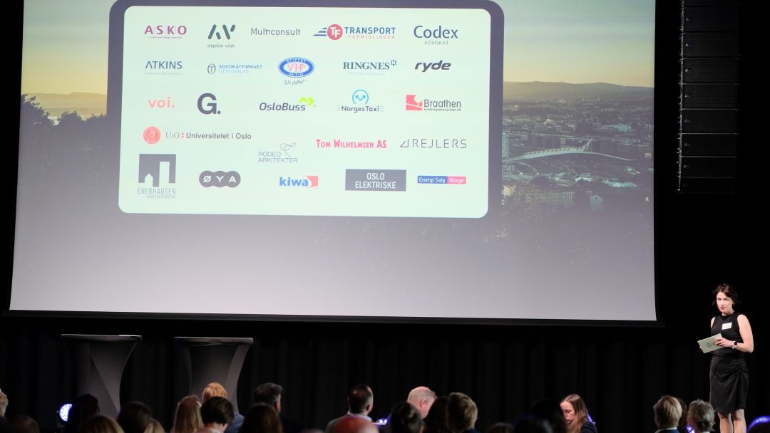 En person står på scenen ved siden av en stor skjerm som viser ulike firmalogoer. Et publikum sitter i forgrunnen.
