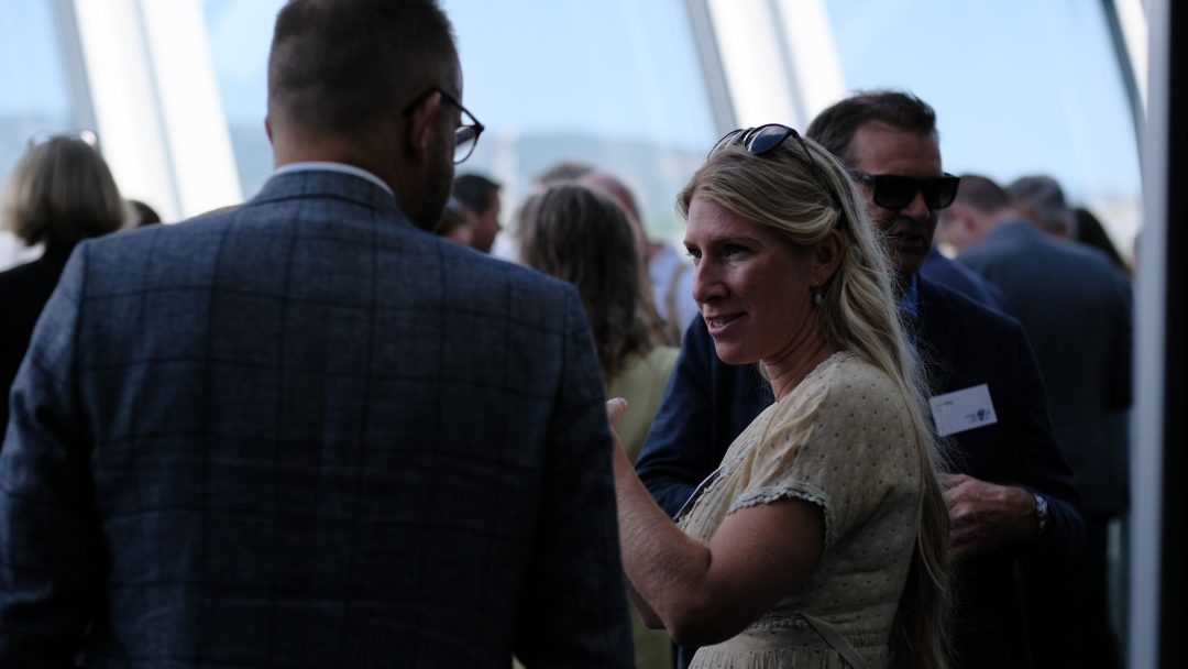 En kvinne i lett kjole snakker med en mann i dress på en innendørs sammenkomst med folk i bakgrunnen.