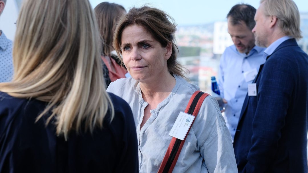 En kvinne med navneskilt og rødstripet veskerem lytter til en annen kvinne. Flere personer snakker utendørs i bakgrunnen.