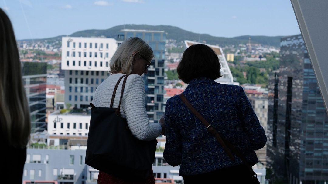 To personer står innendørs og ser ut et vindu på et bybilde med bygninger og åser i bakgrunnen.