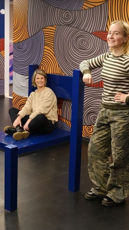 To personer samhandler med overdimensjonerte møbler i et rom med fargerike, virvlende veggmønstre. Den ene sitter på en gigantisk stol fra Oslotips, mens den andre står ved siden av og undrer seg over designet.