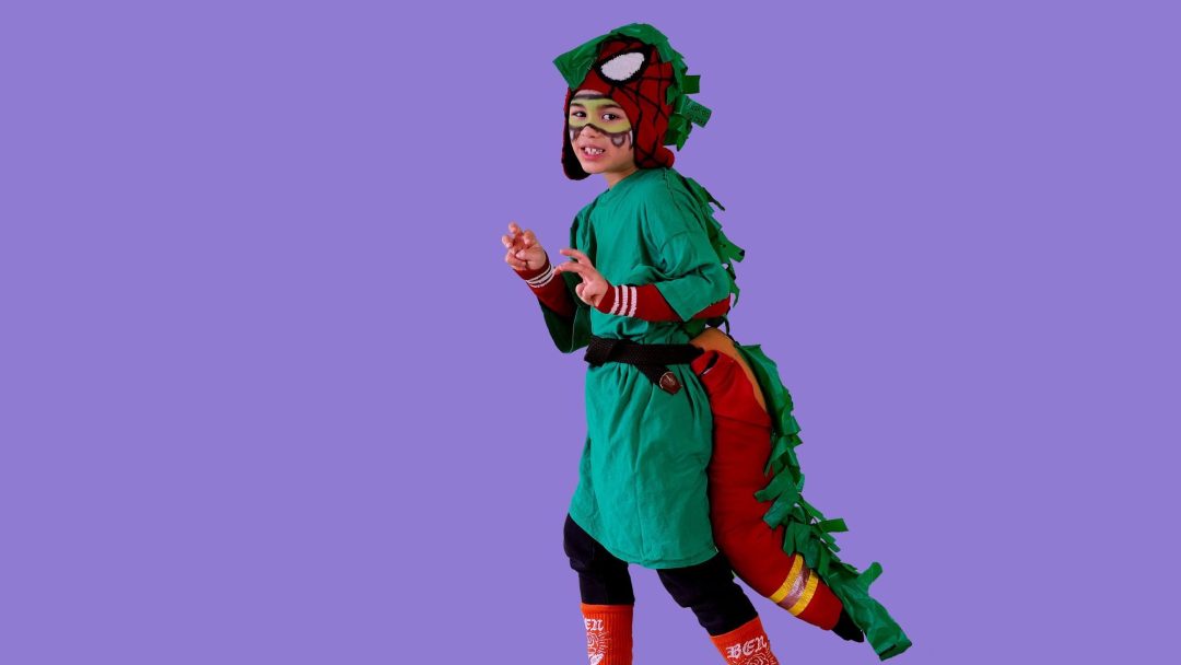 Et barn i et halloweenkostyme-dinosaurantrekk med en grønn kropp, skjell langs ryggen og en lang hale står mot en lilla bakgrunn.