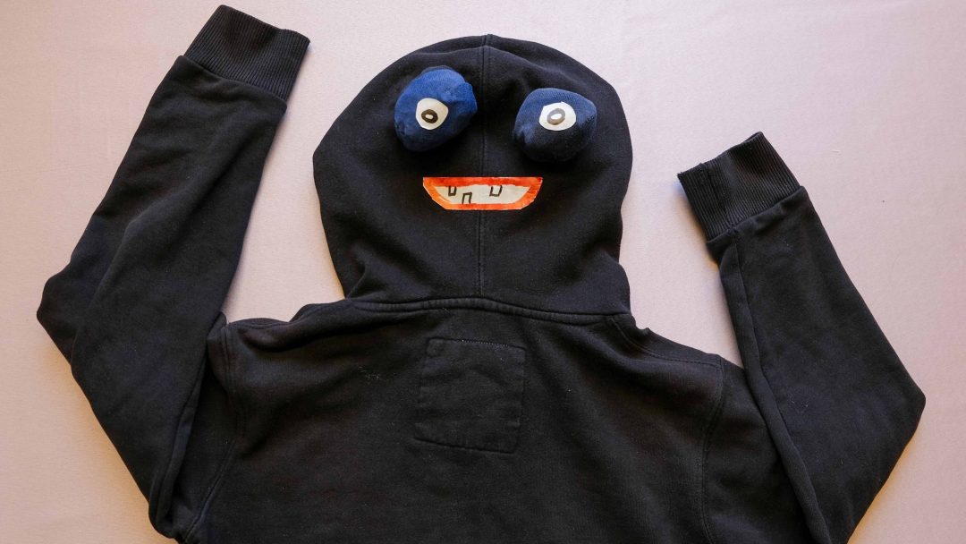 En svart hettegenser med et tegneserielignende ansikt på hetten, med lune øyne og en smilende munn – perfekt for en morsom halloweenkostyme. Armene er plassert oppover på en flat overflate.