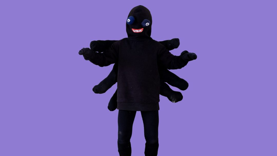 En person kledd i en svart edderkopp halloweenkostyme, komplett med flere utstoppede ben og en smilende maske, står mot en vanlig lilla bakgrunn. Perfekt for å sette et uhyggelig preg på enhver festlig feiring.