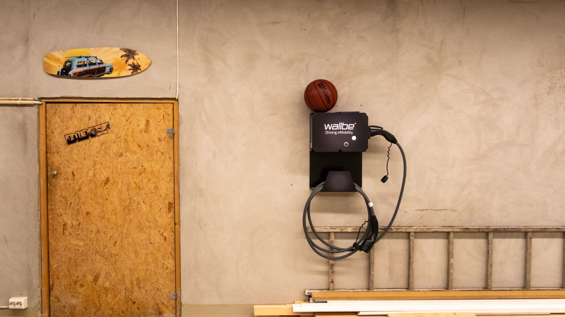 En veggmontert elektrisk kjøretøylader holder en basketball på toppen, flankert av en dør utsmykket med klistremerker og en surfebrettdekor på venstre side. I nærheten venter en almoped tålmodig på sitt neste eventyr langs kystveiene.