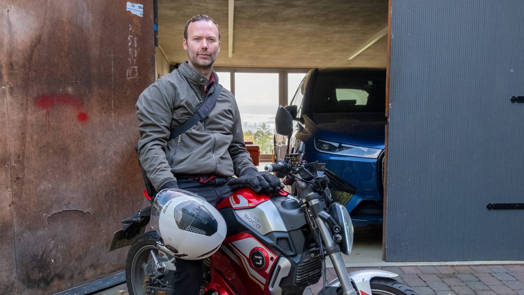 En mann i jakke holder en hjelm, sittende på en elegant rød og sølv elmoped foran en åpen garasje med en bil inni.