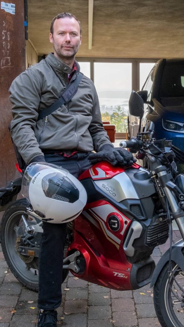 En person sitter selvsikkert på en rød elmoped, holder en hjelm, omgitt av den stille summingen fra parkeringsplassen med en bil parkert i bakgrunnen.