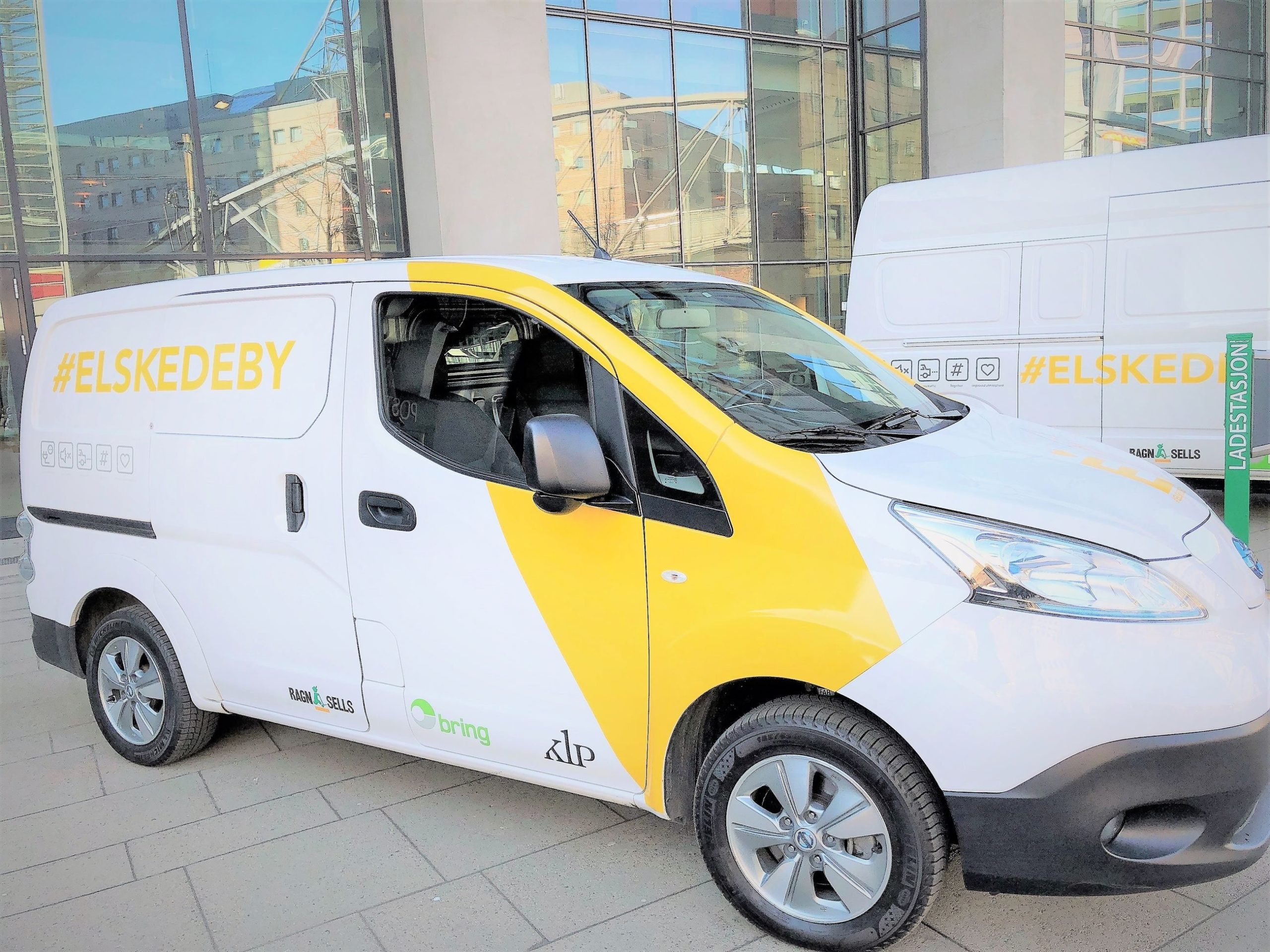 Hvit og gul varebil med emneknaggen #ELSKEDEBY på siden, parkert i et urbant område ved siden av en bygning med glassvinduer.