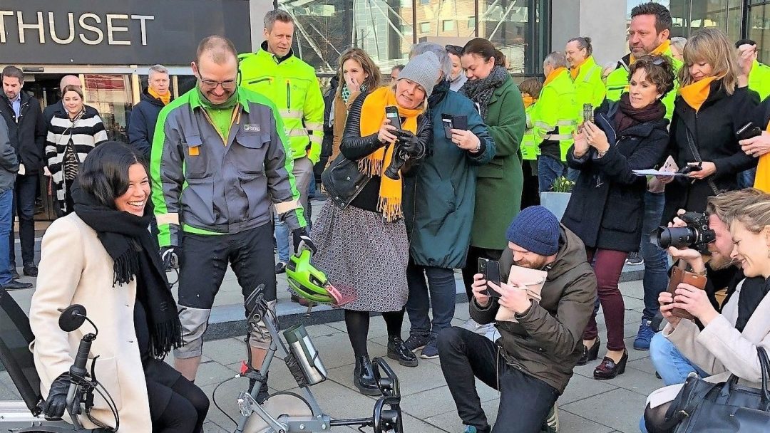 En kvinne sitter på en sykkel omgitt av en gruppe mennesker, noen i synlige jakker, og tar bilder og observerer.