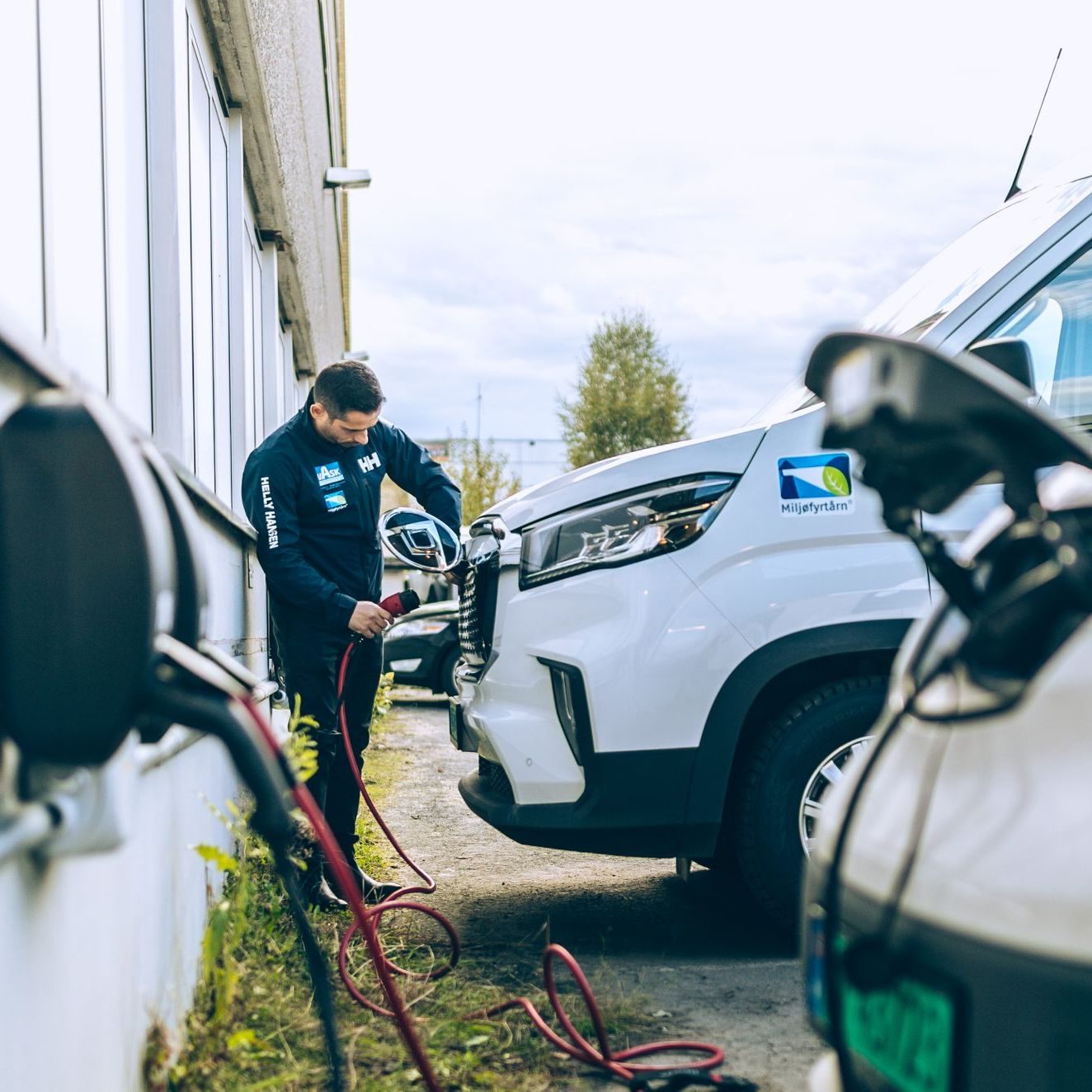 En person kobler en ladekabel til en hvit elektrisk varebil parkert ved siden av et bygg, noe som gjenspeiler initiativene til KlimaOslo. Et annet elektrisk kjøretøy står parkert i forgrunnen med ladekabelen synlig.