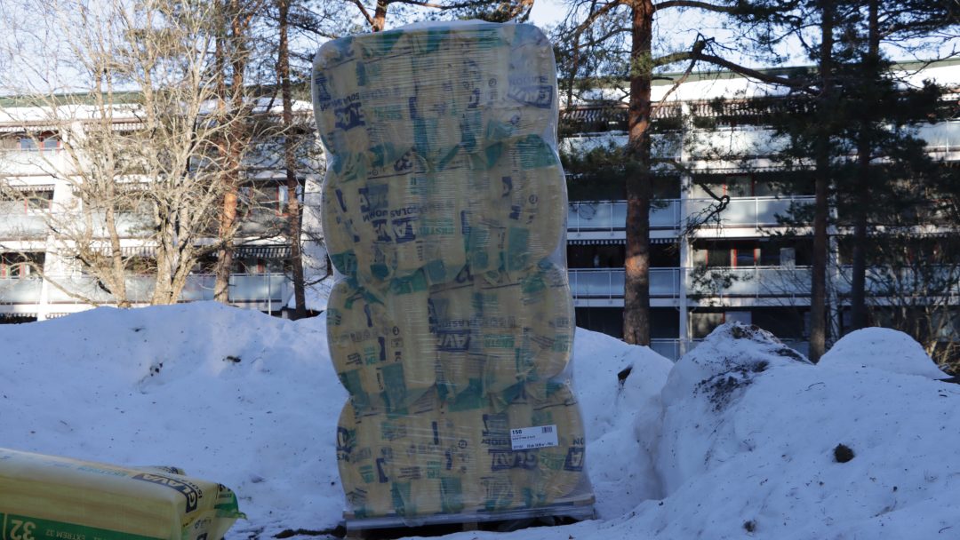 En innpakket pall med isolasjonsmateriale står oppreist på snødekt grunn, støttet av Støtte fra Enova, med trær og en bygning i bakgrunnen.