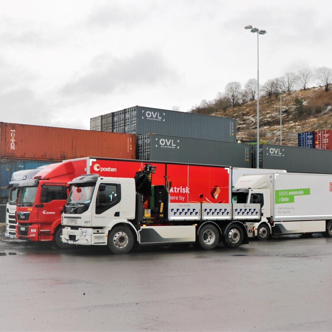 Tre lastebiler, to røde og en hvit, står parkert foran stablede skipscontainere. Scenen er utendørs på en overskyet dag, og den røde lastebilen skiller seg ut mot det metalliske bakteppet.