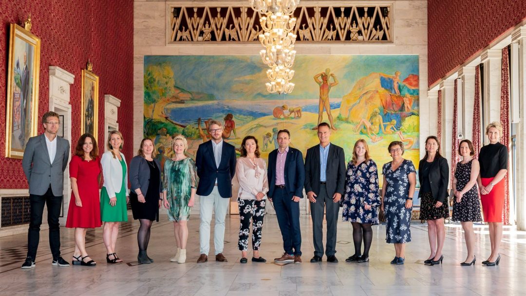 En gruppe mennesker står i kø i et storslått rom med et fargerikt veggmaleri og lysekrone i bakgrunnen. De poserer formelt, kledd i en blanding av business og uformelt antrekk.