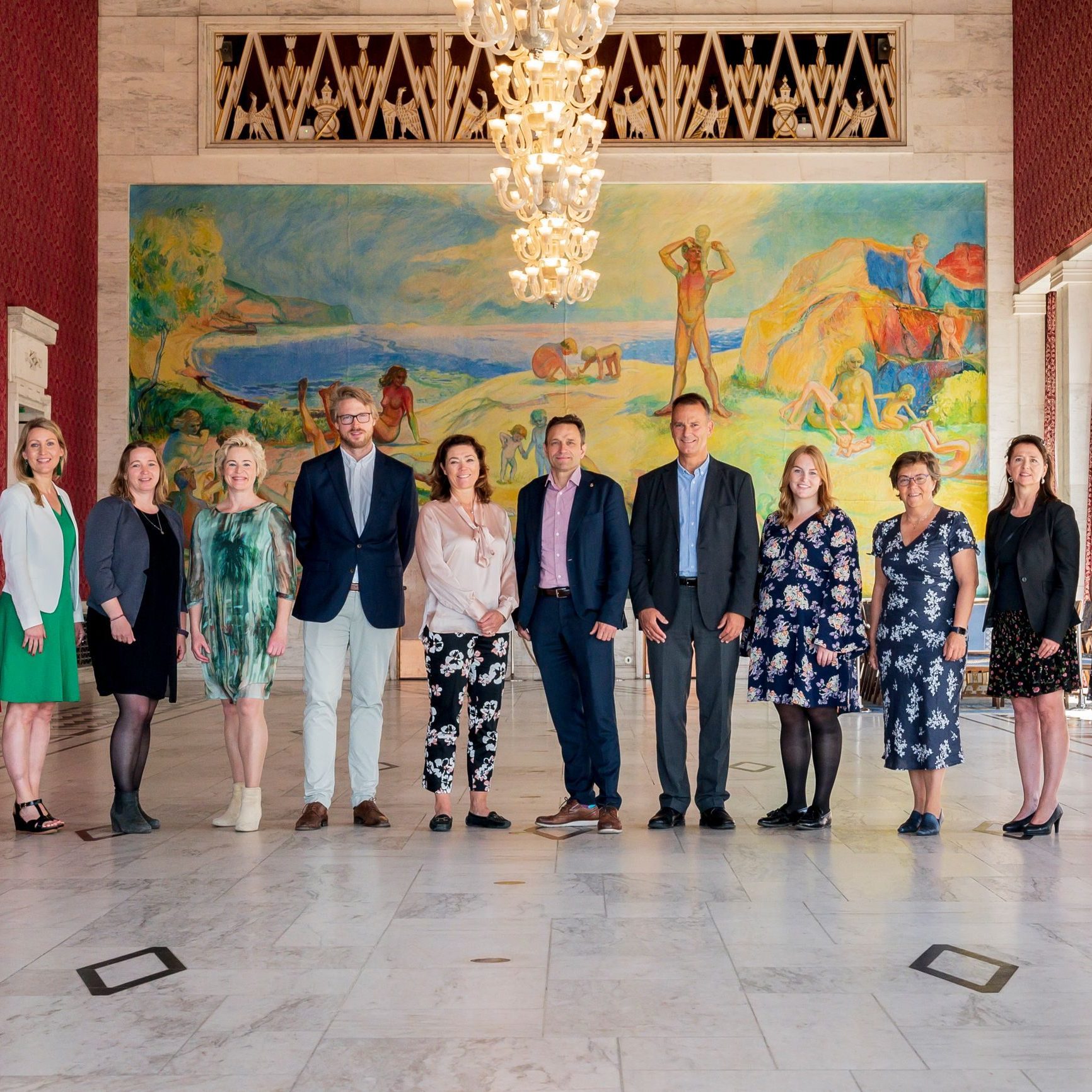 En gruppe mennesker står i et rom med marmorgulv med et fargerikt veggmaleri og lysekrone i bakgrunnen.