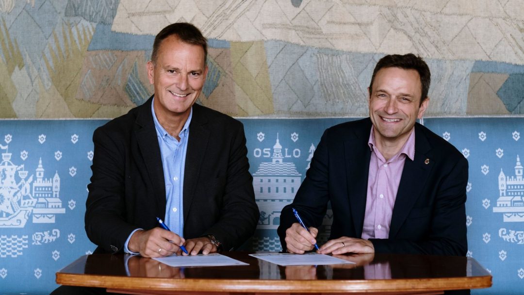 To menn i dress sitter side om side ved et bord, smiler og signerer dokumenter. Et billedvev med geometriske mønstre og ordet OSLO er i bakgrunnen.