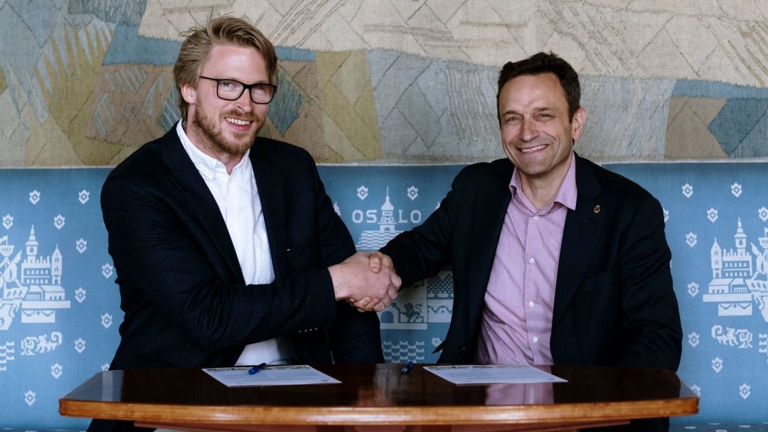 To menn i dress sitter ved et bord, håndhilser og smiler til kameraet. Dokumenter og penner ligger på bordet foran dem.