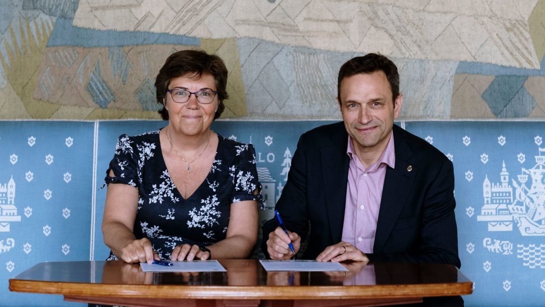 To personer som sitter ved et bord og signerer dokumenter med penner, foran et mønstret bakteppe.