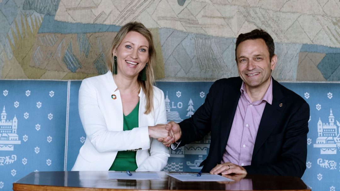 To personer som sitter ved et bord, håndhilser og smiler, med dokumenter foran seg. De står i et rom med mønstret tapet.