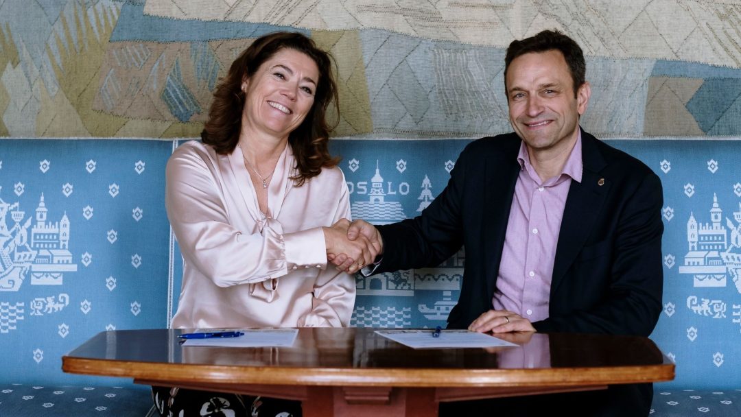 To personer, en kvinne og en mann, sitter ved et bord, smiler og håndhilser. Et stykke papir og penn er synlig på bordet. Bakgrunnen har et geometrisk mønster.