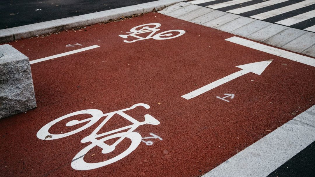 Den røde sykkelveien med hvite sykkelsymboler og en pil viser tydelig hvilke sykkelregler syklister må følge, mens den går langs et fotgjengerovergang med sebrastriper.