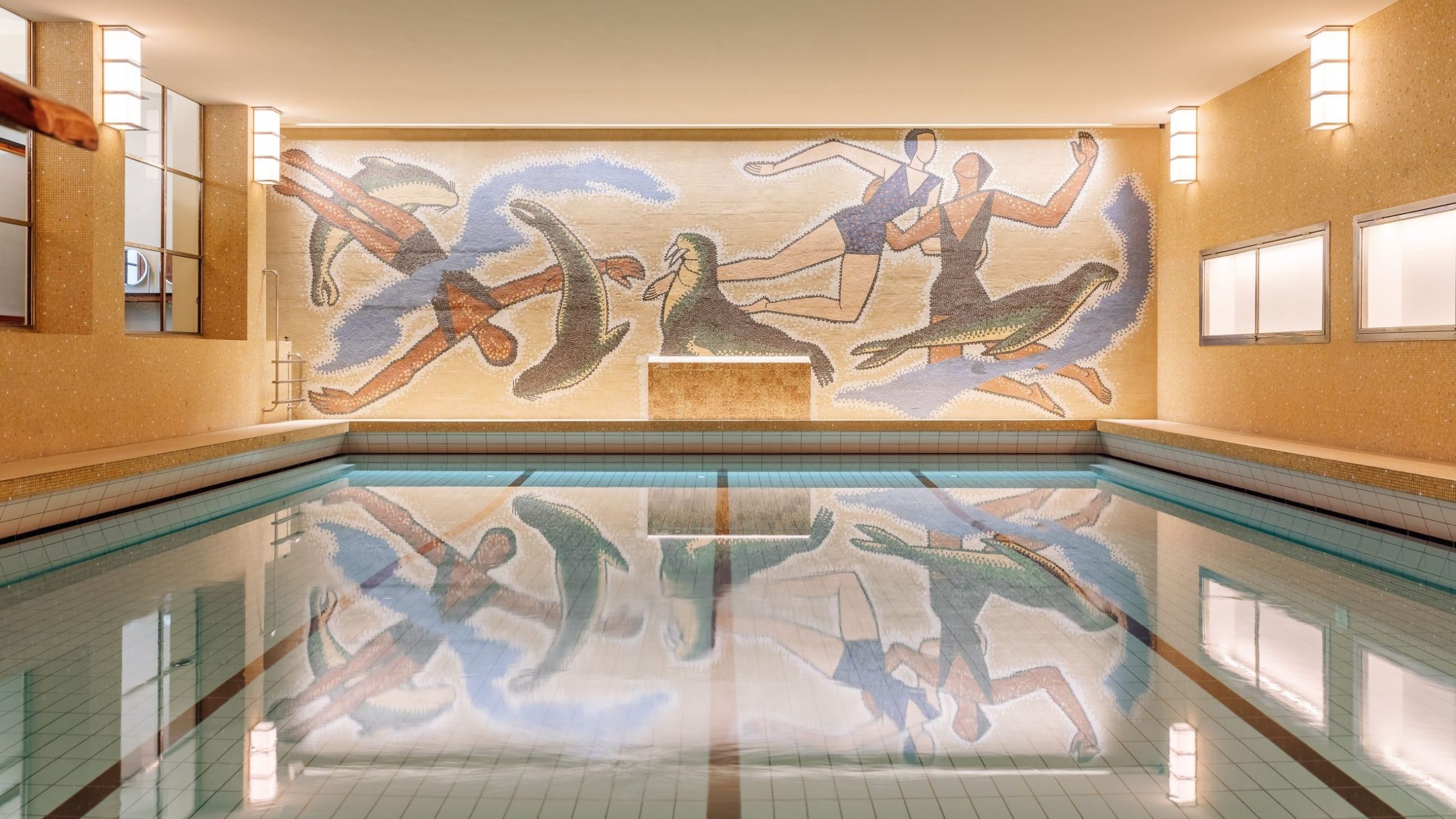 Innendørsbassenget har flislagt gulv og beige vegger, fremhevet av et fargerikt veggmaleri av abstrakte menneskefigurer og fugler på den ytterste veggen. Store overlys lyser opp rommet og skaper en innbydende atmosfære som minner om populære Oslotips for avslapning og fritid.