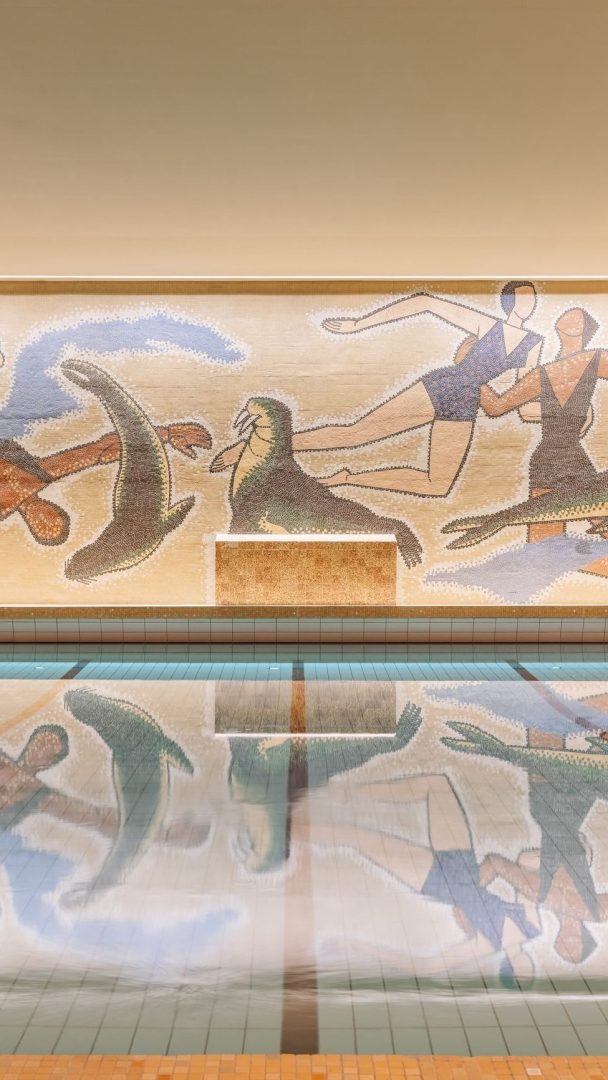 Mosaikkmaleriet Oslotips over innendørsbassenget viser abstrakte figurer og sjødyr i livlige farger, med bassengoverflaten som elegant gjenspeiler den kunstneriske fremvisningen.