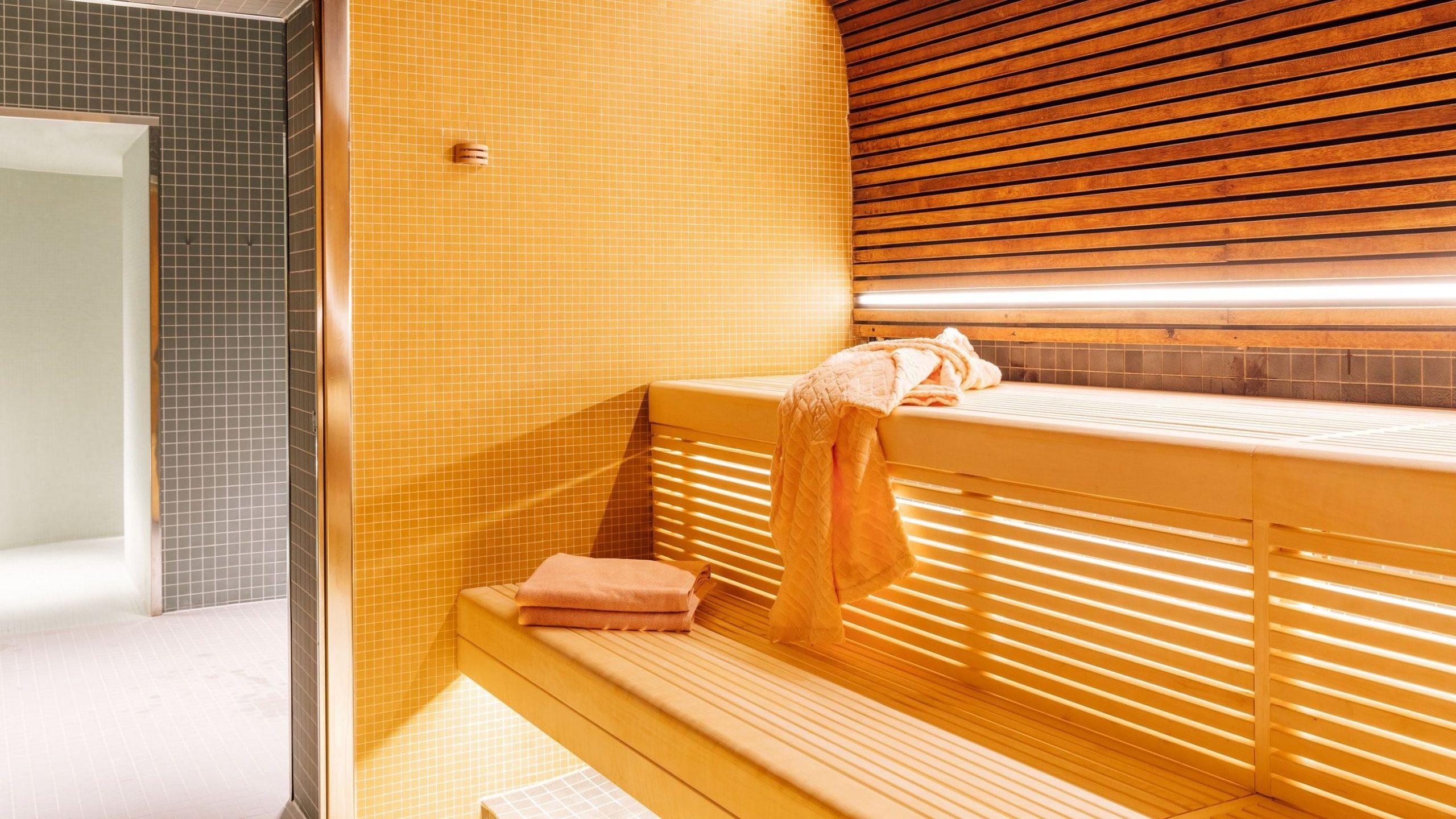 Flislagt badstue med trebenk og spaltevegger, med brettede håndklær. Kraftig belysning over og en åpen dør som leder til et tilstøtende flislagt rom, og tilbyr en ekte Oslotips-opplevelse.