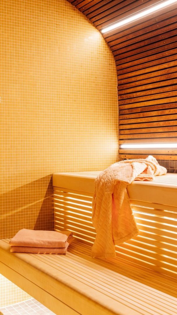 Badstuinteriøret i tre viser varm, gylden belysning og håndklær pent plassert på benker. Vegger utsmykket med et lysegult mosaikkmønster fullfører dette rolige rommet, og tilbyr et tilfluktssted som er verdt Oslotips for avslapning.